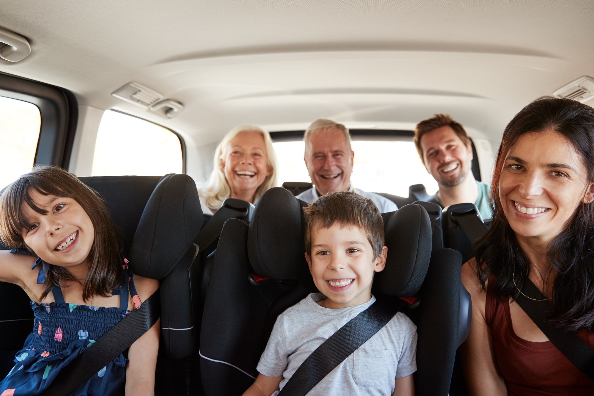 Three generation white family sitting in two rows of passenger seats in a car, smiling to camera